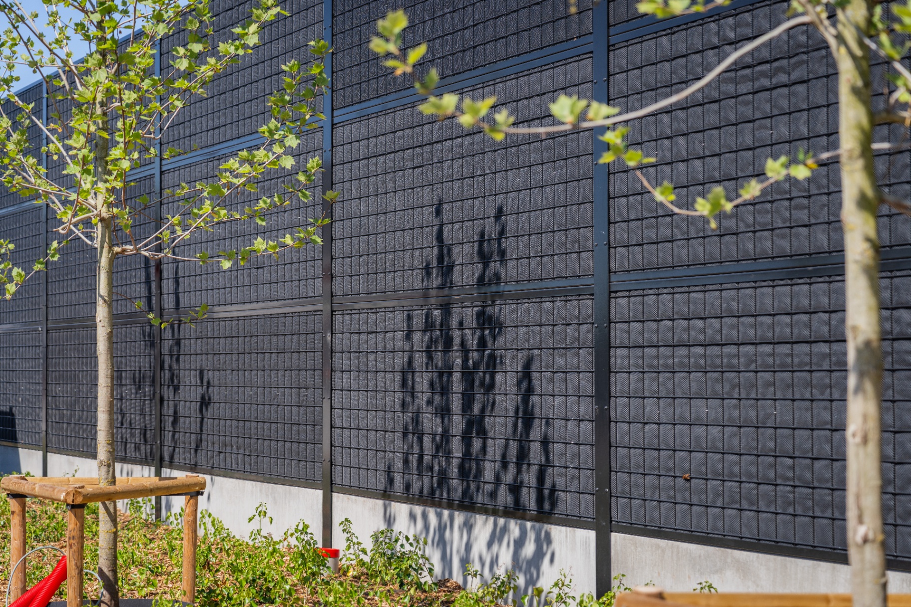 Tall Noise-Reducer wall at Leievoeders with young trees in front, shielding noise from the neighbours