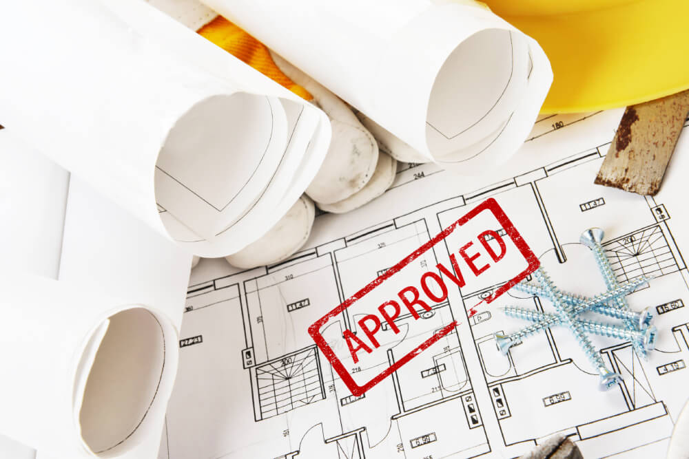 Goedgekeurde bouwplannen met "APPROVED"-stempel, opgerolde tekeningen, veiligheidshelm en schroeven op de tekentafel
