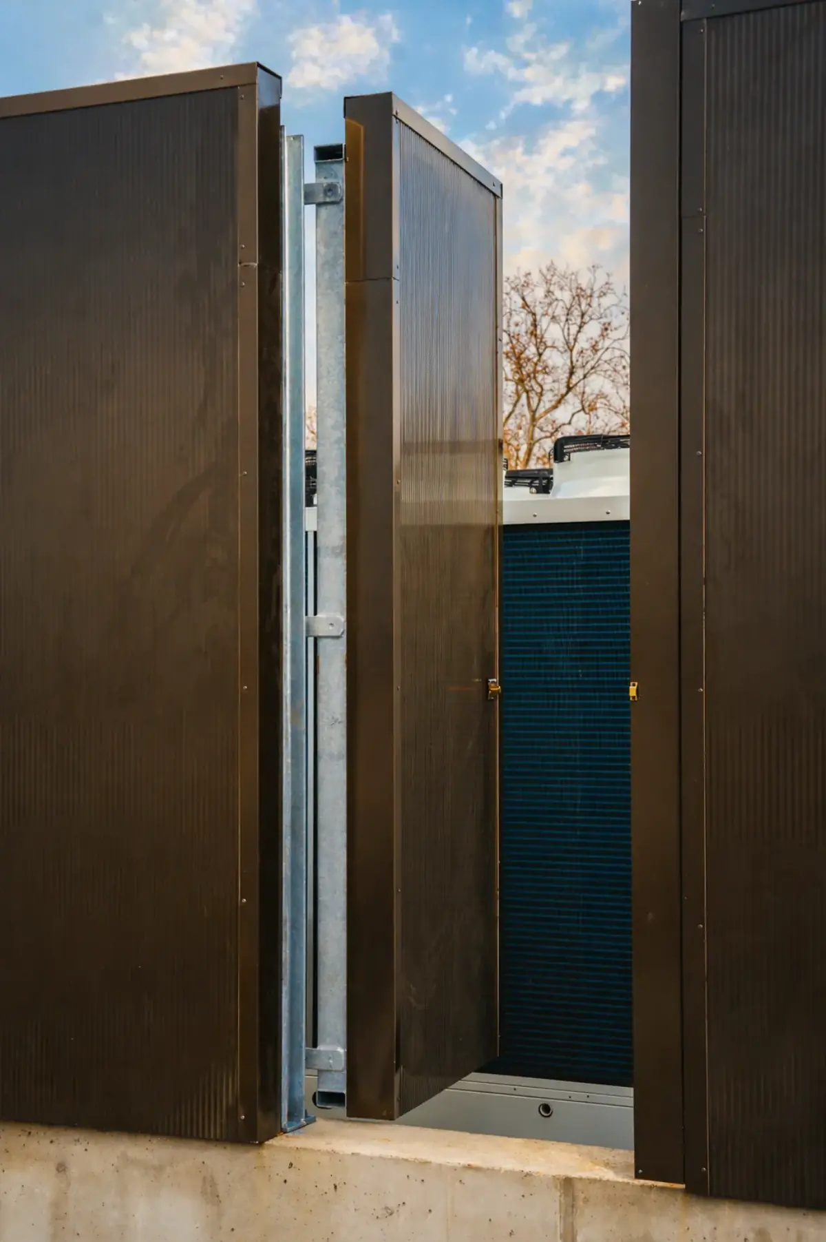 Détail d'un panneau Ichono Wall avec porte d'accès ouverte, montrant les tôles d'acier profilées sombres avec l'installation technique et un élément filtrant bleu en arrière-plan