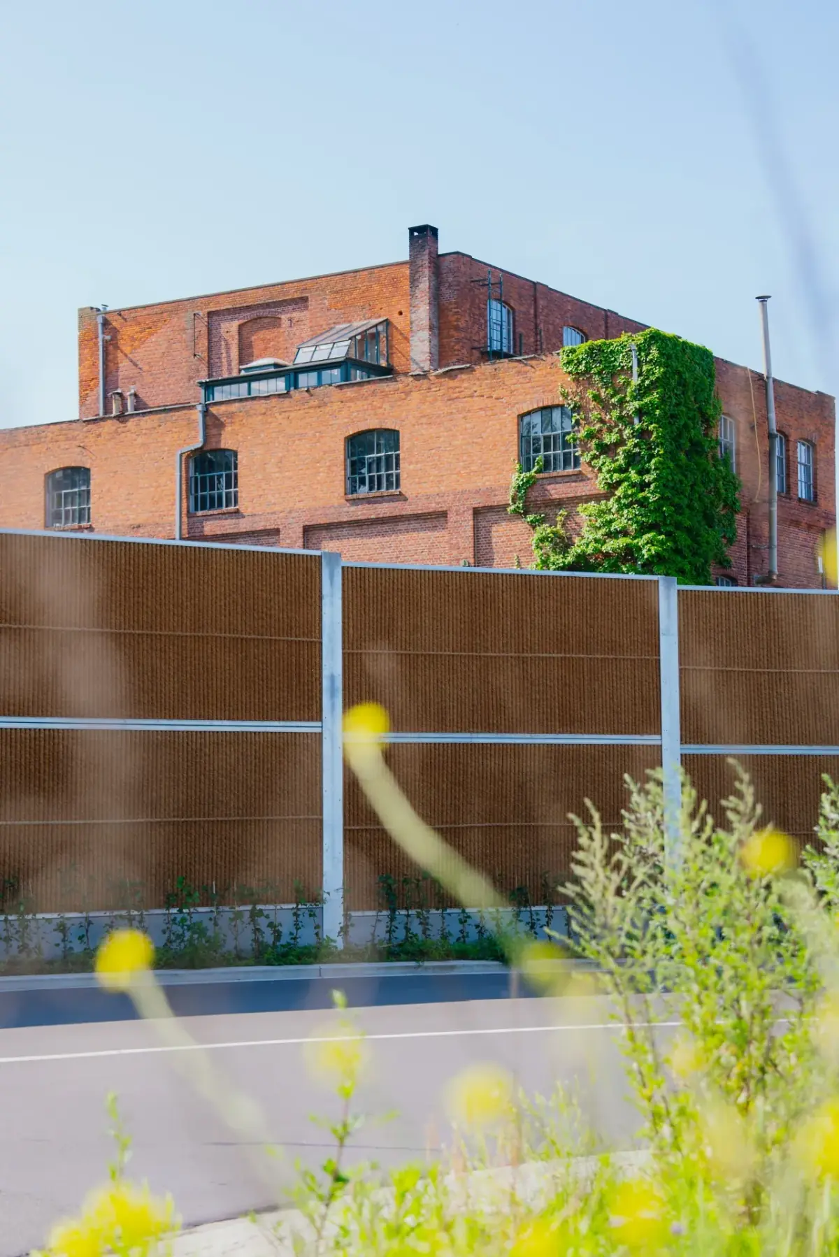 Kokowall sound barrier along a road with a natural coconut fibre finish and a brick industrial building in the background